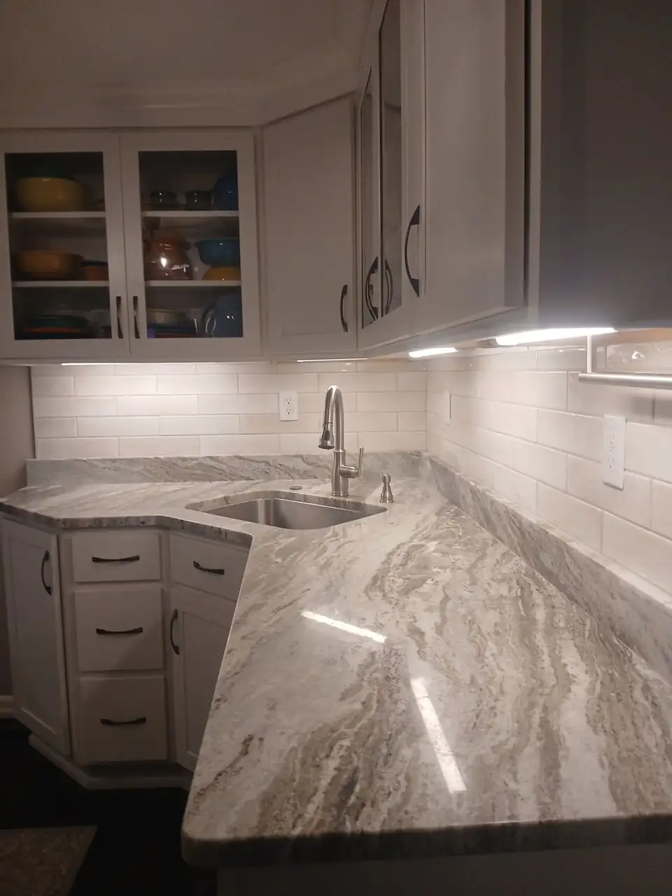 Kitchen by a remodeling contractor in Erie: white cabinets with glass doors, a flowing gray and taupe stone countertop, undermount sink and white subway tile backsplash