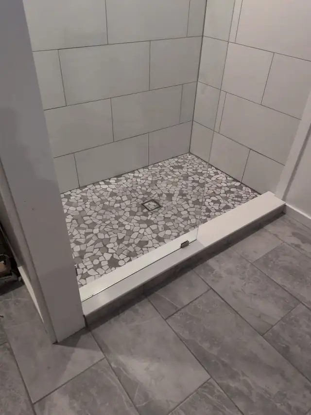 Walk-in shower with pebble mosaic floor, square drain, large white wall tiles, white curb, glass panel and gray stone-look floor tile