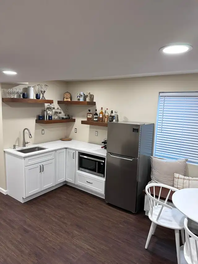 White kitchenette with floating wood shelves, white countertop, stainless refrigerator and microwave